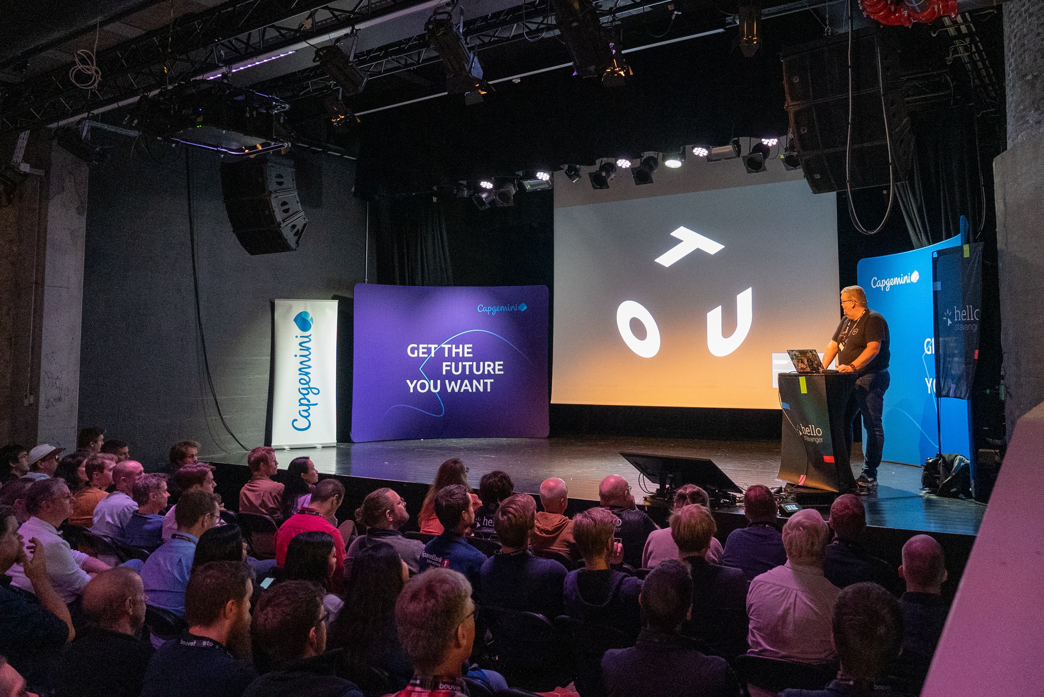 A speaker at the lectern addressing a seated audience beneath the Tou logo.